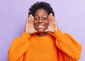 femme-garde-paumes-pres-du-visage-sourit-largement-yeux-pleins-bonheur-idiots-autour-porte-pull-tricot-orange-isole-violet-freepik