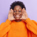 femme-garde-paumes-pres-du-visage-sourit-largement-yeux-pleins-bonheur-idiots-autour-porte-pull-tricot-orange-isole-violet-freepik